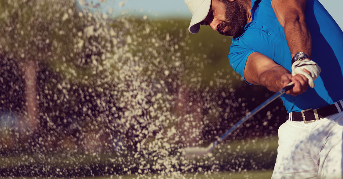 golfer in blue shirt and white hat hits ball out of sand trap